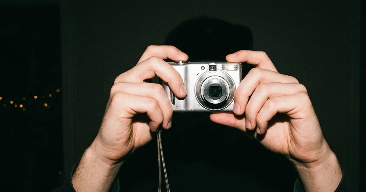 Two hands holding a small silver vintage compact camera toward the viewer in a dark room, lit by a bright direct flash